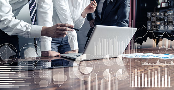 Gruppen von 3 Personen steht in einem Konferenzraum vor einem Notebook. Im Vordergrund schweben schwach eingeblendet statistische Auswertungen im digitalen Raum.